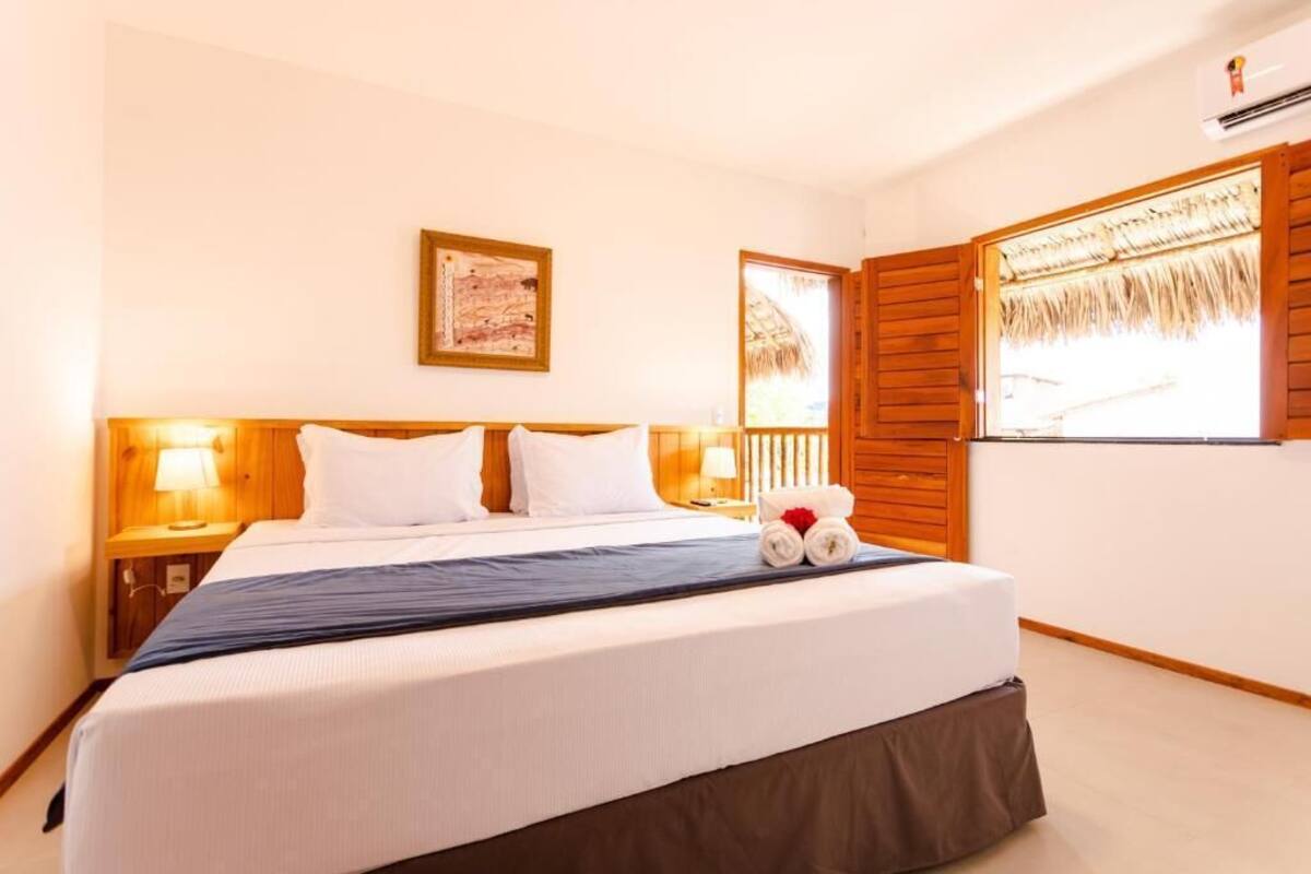 Habitación de hotel con una cama grande, cabecera de madera, dos lámparas de noche, un cuadro enmarcado en la pared y una ventana con contraventanas de madera.