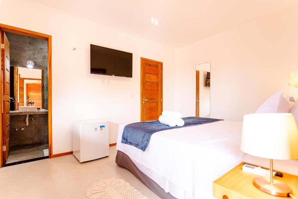 A hotel room with a bed, a TV mounted on the wall, a small refrigerator, and a bedside table with a lamp.