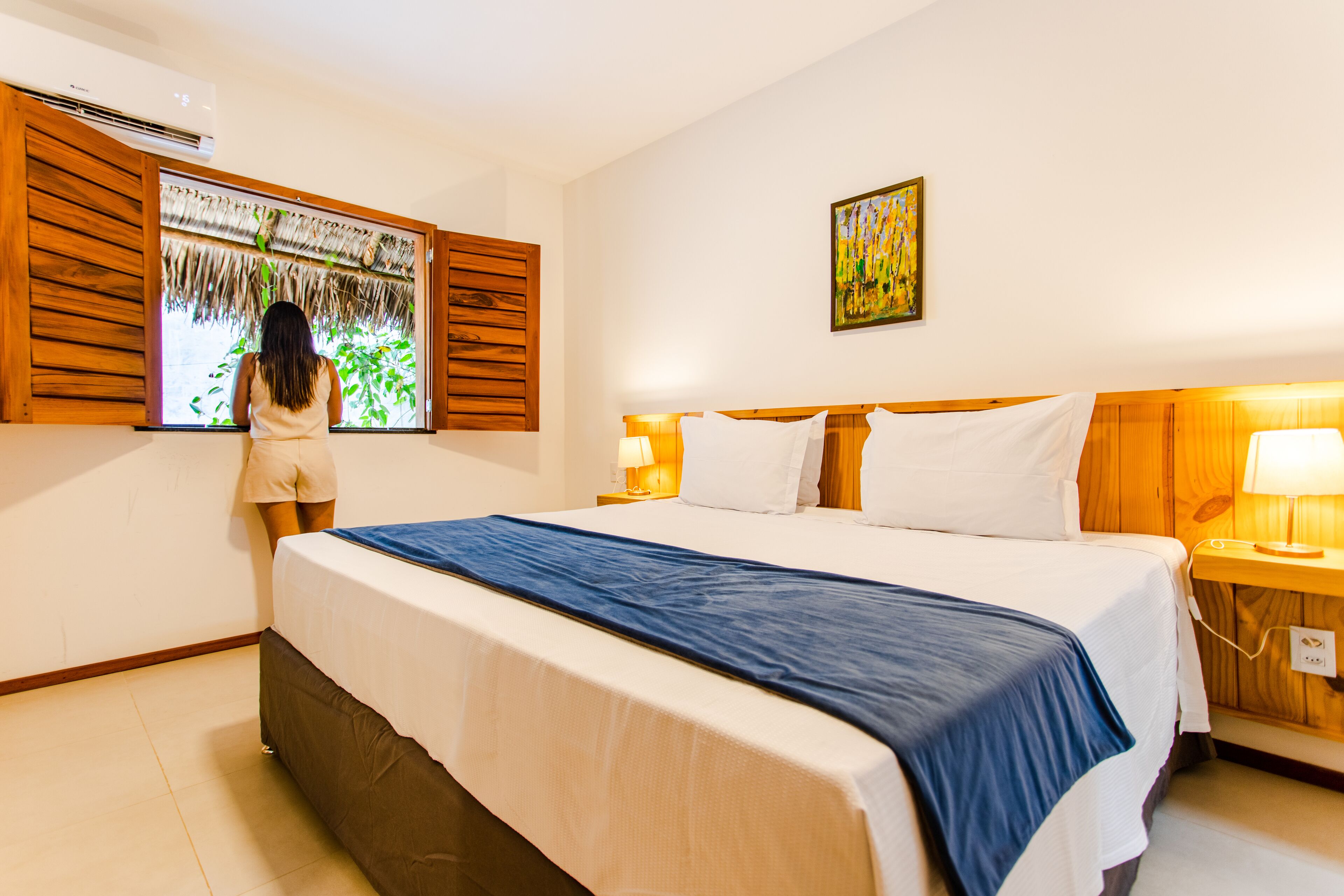 Quarto de hotel com uma cama grande, cabeceira de madeira, janela com vista para o verde e um quadro na parede.