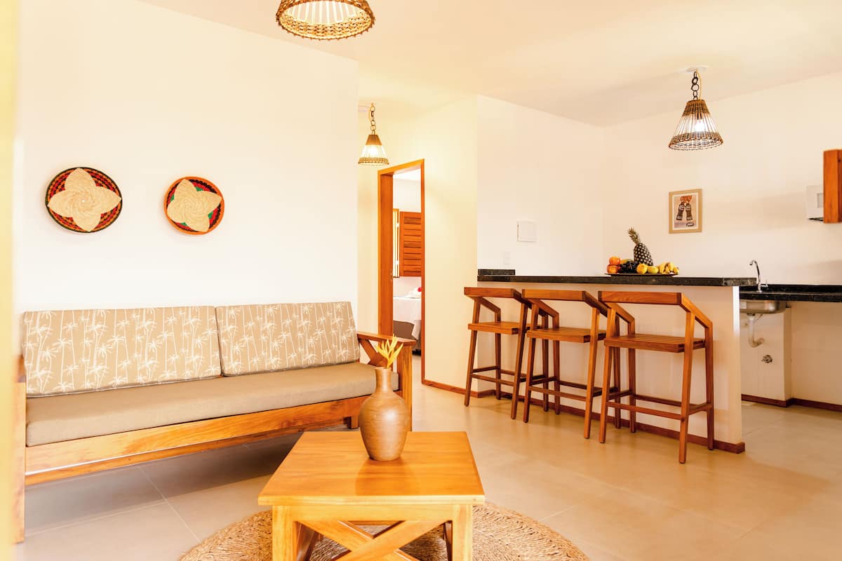 Una sala de estar moderna con un sofá de madera, una pequeña mesa con un jarrón y una cocina con taburetes de madera y una encimera negra.