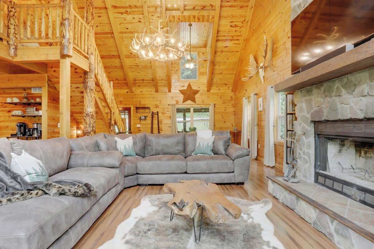 A log cabin interior with a large gray sectional sofa, a stone fireplace, and a wooden ceiling.