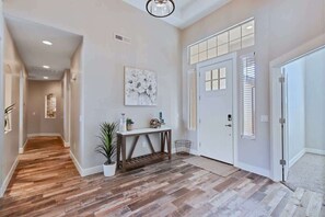 Un pasillo con suelo de madera, una puerta blanca con persianas, una consola decorada con objetos y una planta en maceta.
