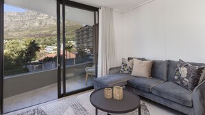 A modern living room with a grey sofa, two cushions, a small round table with two candle holders, and a view of a mountainous landscape through large glass doors. - The Cedar Unit 105 (Cape Town)