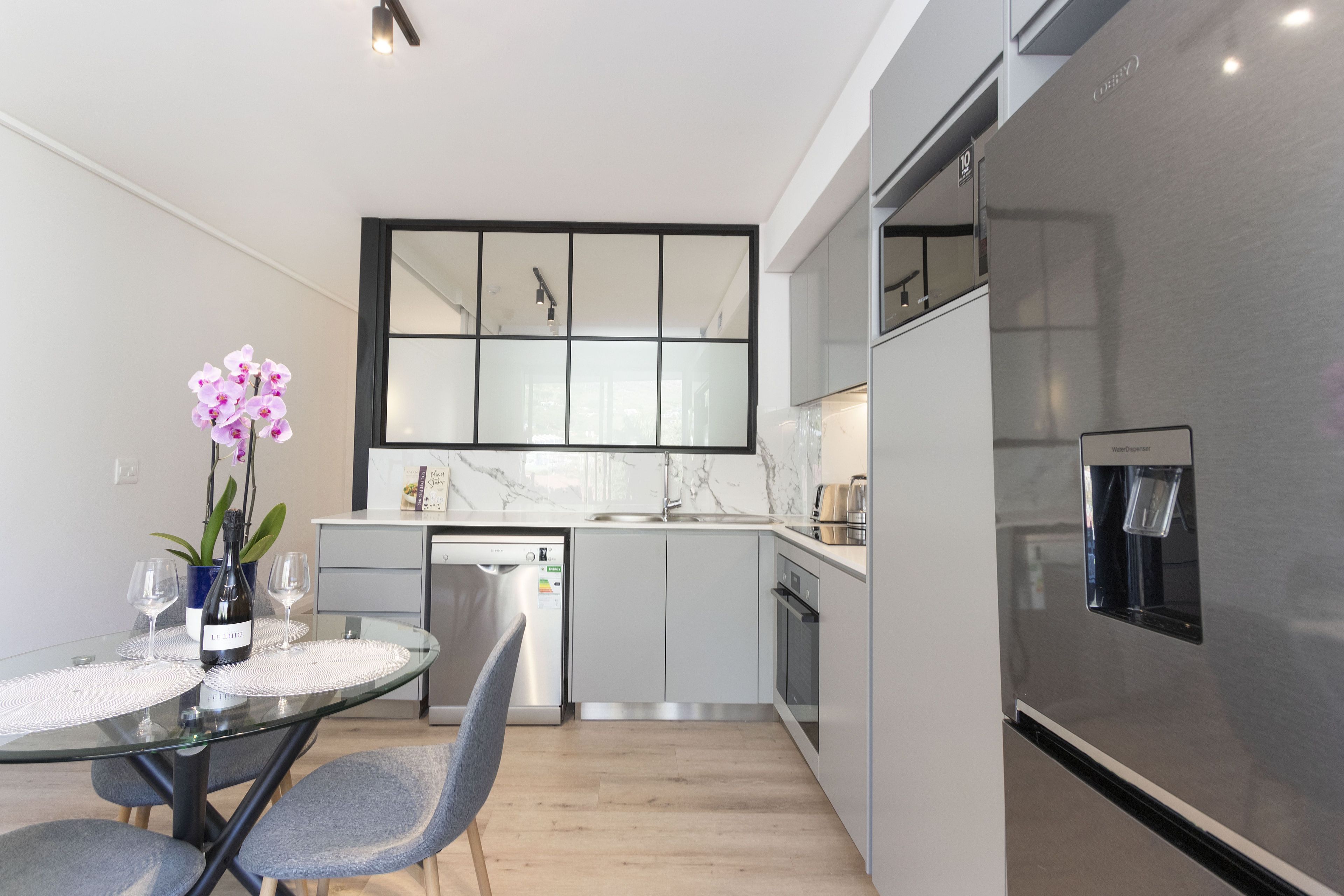 Une cuisine moderne avec une table à manger en verre, des chaises, une bouteille de vin et une orchidée en pot.