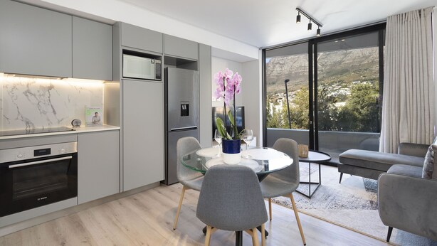A modern kitchen with grey cabinets, a marble backsplash, and a built-in oven. There is a dining area with a glass table and grey chairs, and a large window with a view of a mountainous landscape. - The Cedar Unit 105 (Cape Town)