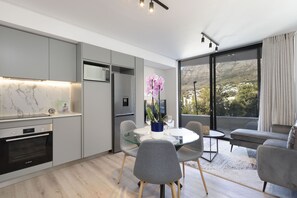 Une cuisine moderne avec des meubles gris, un pare-feu en marbre et un four encastré. On y trouve un coin repas avec une table en verre et des chaises grises, ainsi qu’une grande fenêtre donnant sur un paysage montagneux.