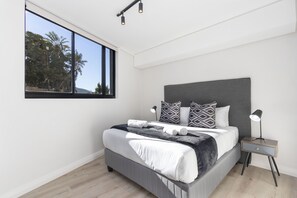 A modern bedroom with a large bed, a nightstand, a desk lamp, and a window with a view of trees.