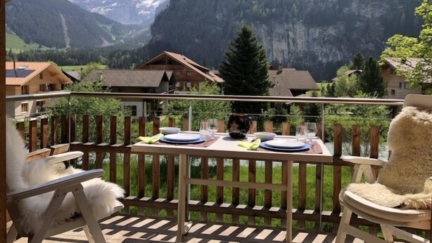 Ein Balkon mit Esstisch, der auf eine Berglandschaft mit Häusern und einem bewaldeten Gebiet blickt.