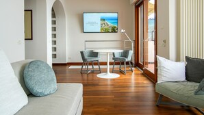 Ein modernes Wohnzimmer mit einer Couch, einem Esstisch mit Stühlen, einem an der Wand montierten Fernseher und einem großen Fenster mit Jalousien.