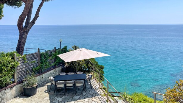 Eine Terrasse mit Tisch und Stühlen, einem großen Sonnenschirm und Blick auf das Meer.