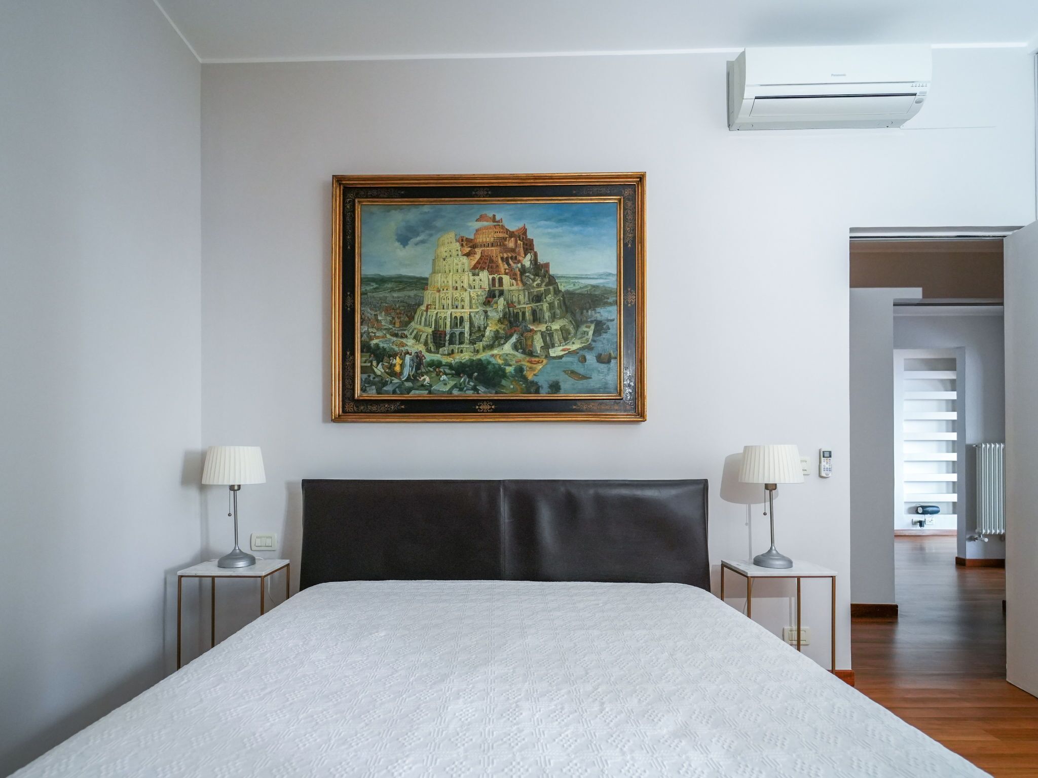 Ein Schlafzimmer mit einem großen Bett, zwei Nachttischen mit Lampen, einem gerahmten Gemälde eines historischen Gebäudes und einer Klimaanlage.