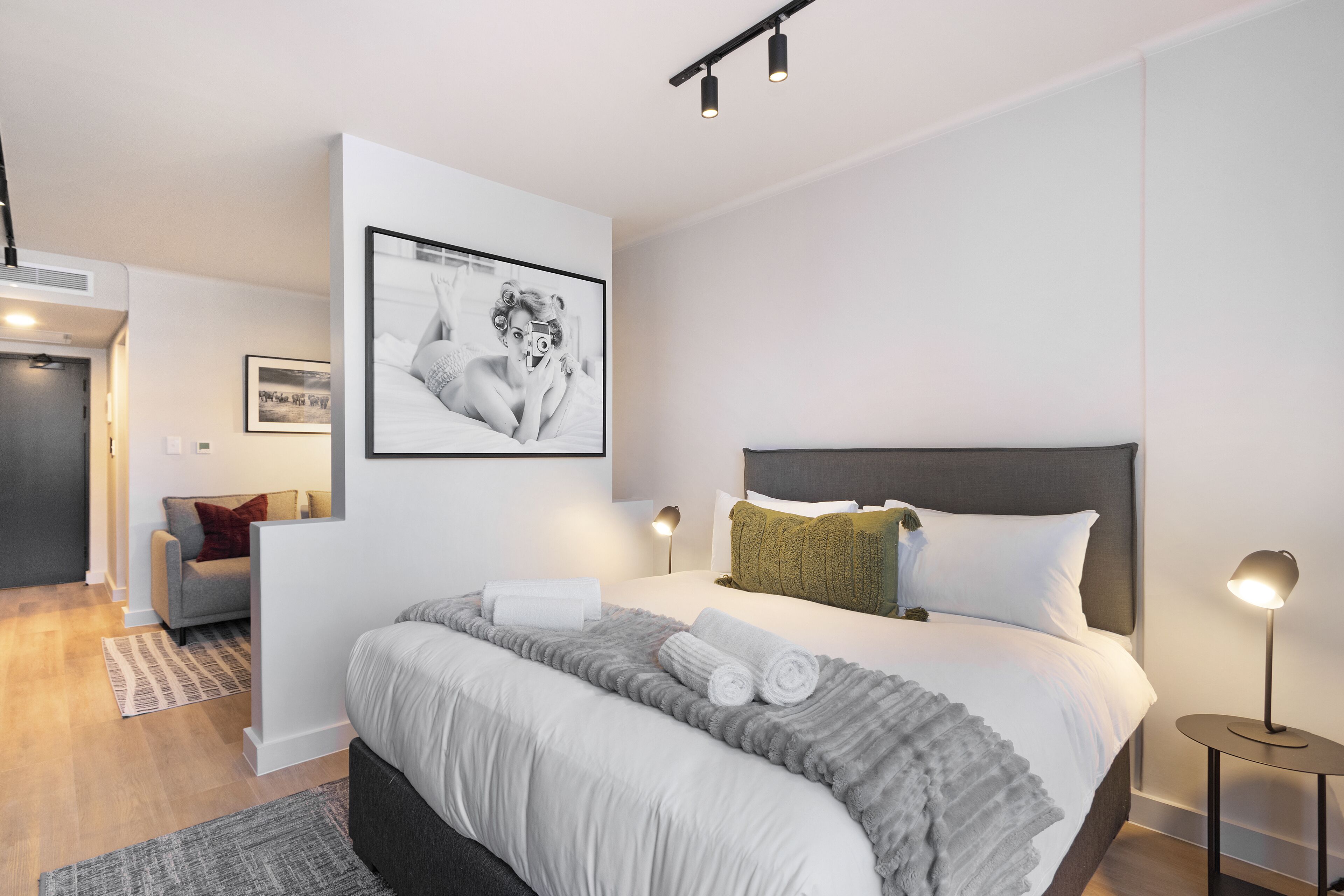 A modern hotel room with a large bed, a grey and white color scheme, a framed photograph on the wall, and a small side table with a lamp.