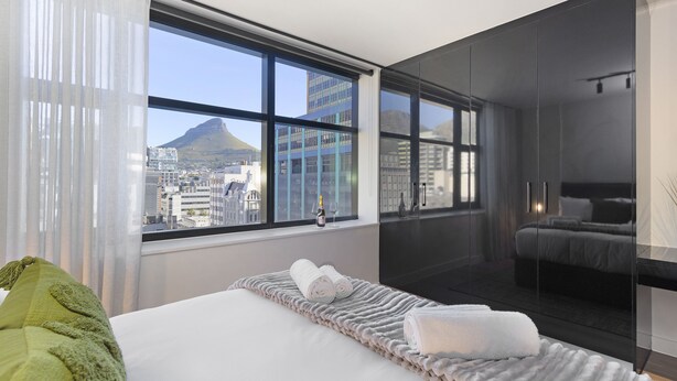 A modern hotel room with a large window offering a city view, a neatly made bed, and a glass-enclosed walk-in closet. - The Tokyo 807 (Cape Town)