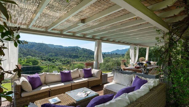 A covered outdoor seating area with wicker furniture, a table, and a view of a mountainous landscape.