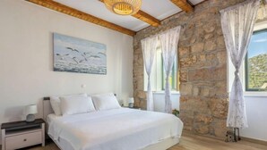 Un dormitorio con pared de piedra, una cama con ropa de cama blanca, una mesita de noche con lámpara y un cuadro de pájaros en la pared.