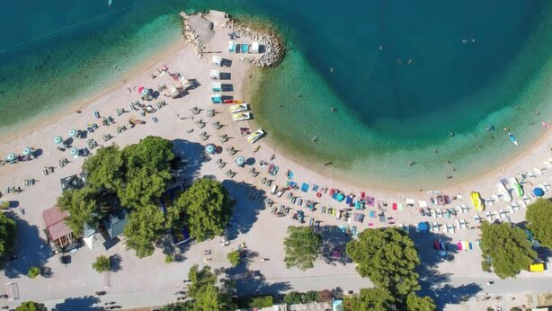 Vista aérea de una playa con filas de tumbonas y sombrillas, una pequeña zona rocosa y aguas azules cristalinas.