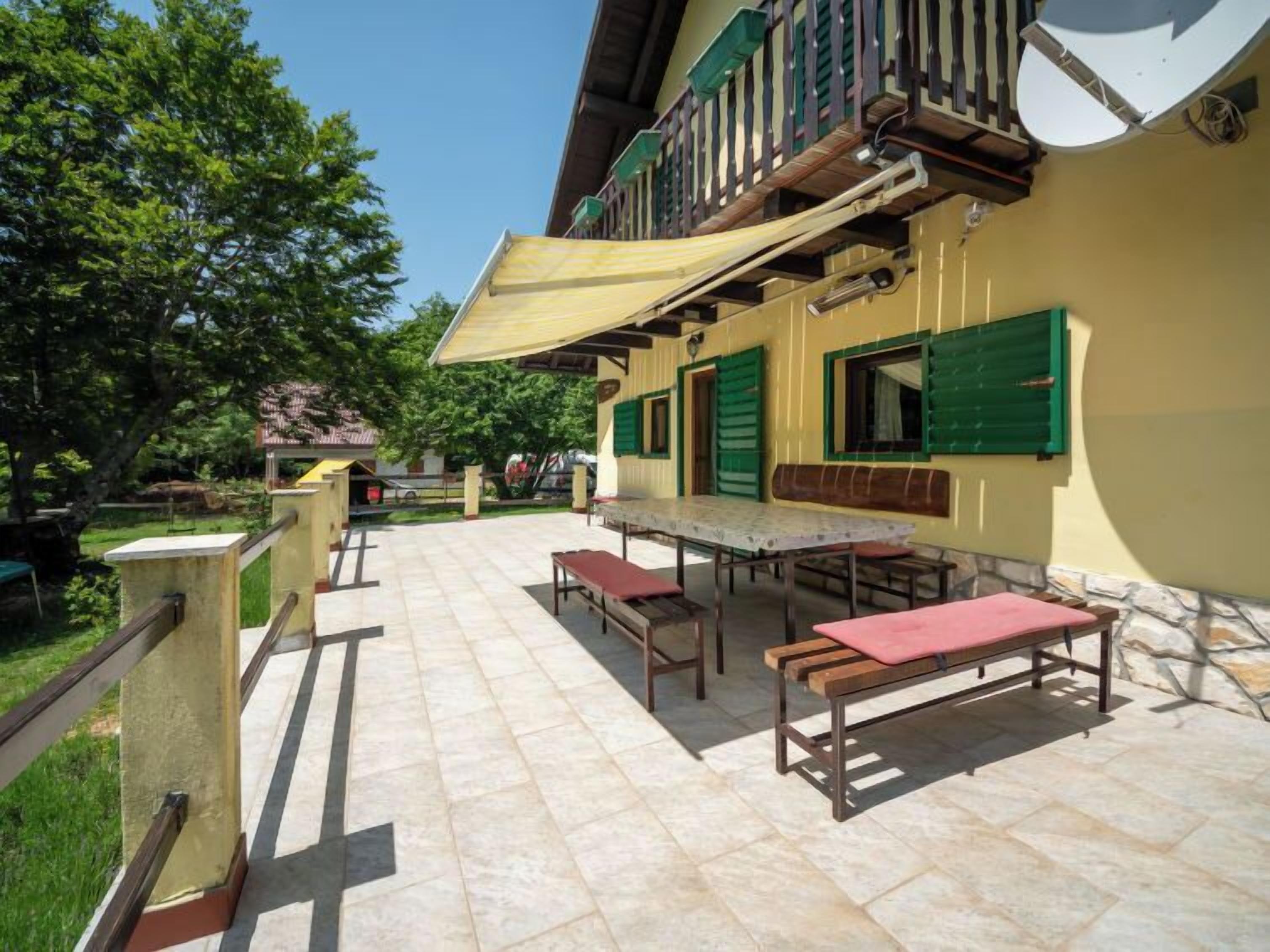 An outdoor patio area with a long table and benches, surrounded by a yellow building with green shutters and a balcony.