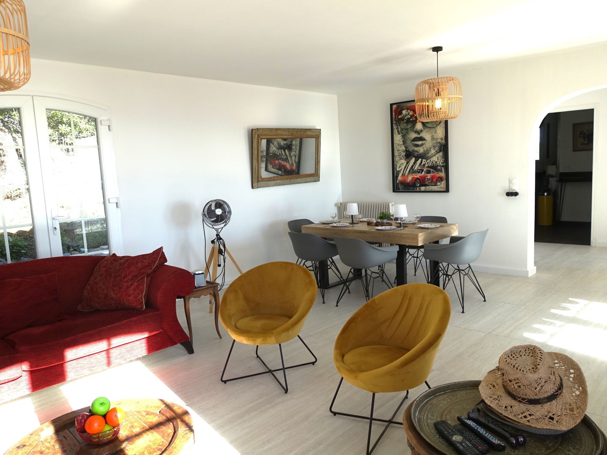 A modern living room with a red sofa, yellow chairs, a dining table with chairs, a fan, and a painting of a car.