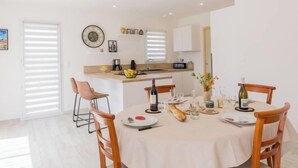Een moderne eetruimte met een ronde tafel voor vier personen, een keuken op de achtergrond en een groot raam met zonwering.