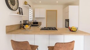 Een moderne keuken met witte kastjes, een houten aanrechtblad en een grote wandklok. Twee bruine stoelen staan bij het aanrecht, er is een schaal met fruit en een fornuis.