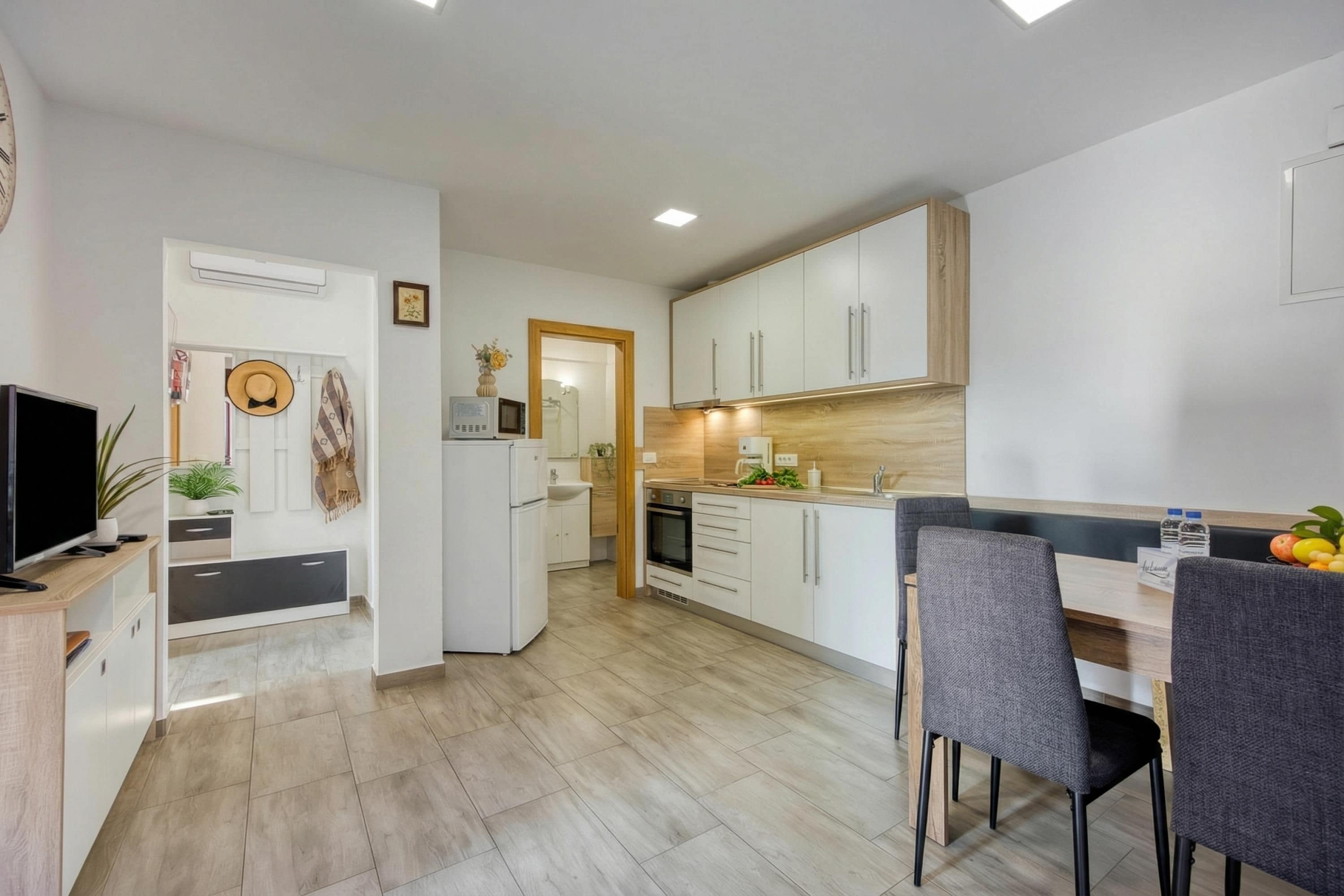A modern kitchen with white cabinets, a dining area with grey chairs, and a TV on a wooden stand.