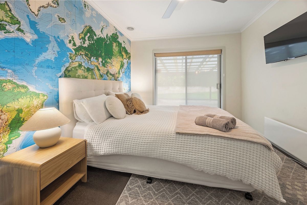 Une chambre avec une grande carte du monde accrochée au mur, un lit recouvert d’une couette blanche, une table de chevet en bois avec une lampe, et un téléviseur fixé au mur.