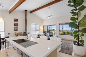 A modern kitchen with a central island, a dining area with a table and chairs, a sofa, and a large window with a view.