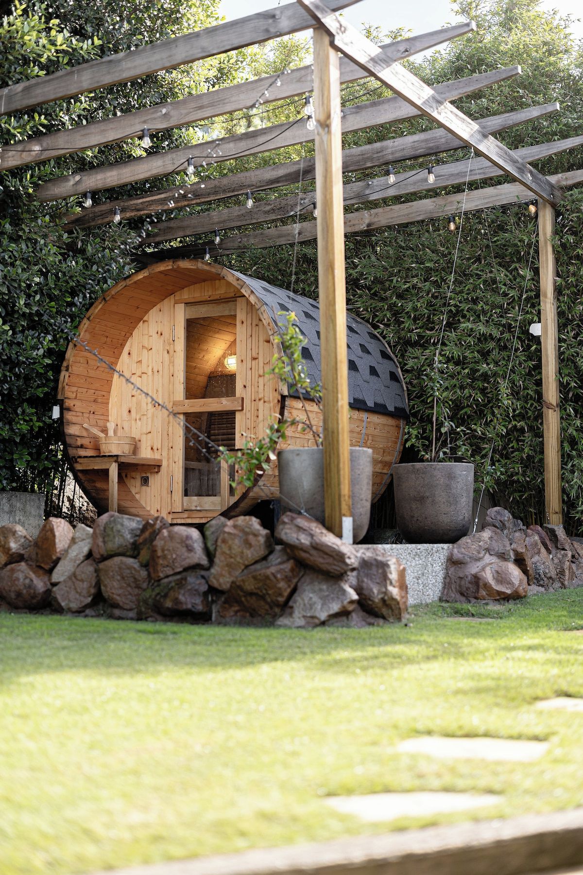 A wooden cabin with a curved roof, a stone wall in front, and a pergola overhead.