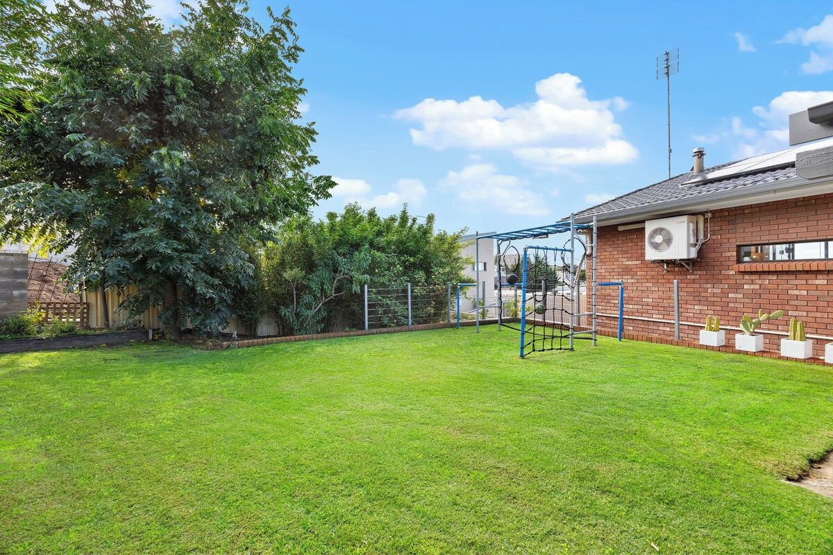 A backyard with a swing set, a brick house, and a large tree.
