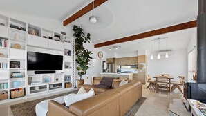 Ein modernes Wohnzimmer mit einem großen Flachbildfernseher, einem Ledersofa, einem Essbereich mit Holztisch und Stühlen sowie einer Küche im Hintergrund.