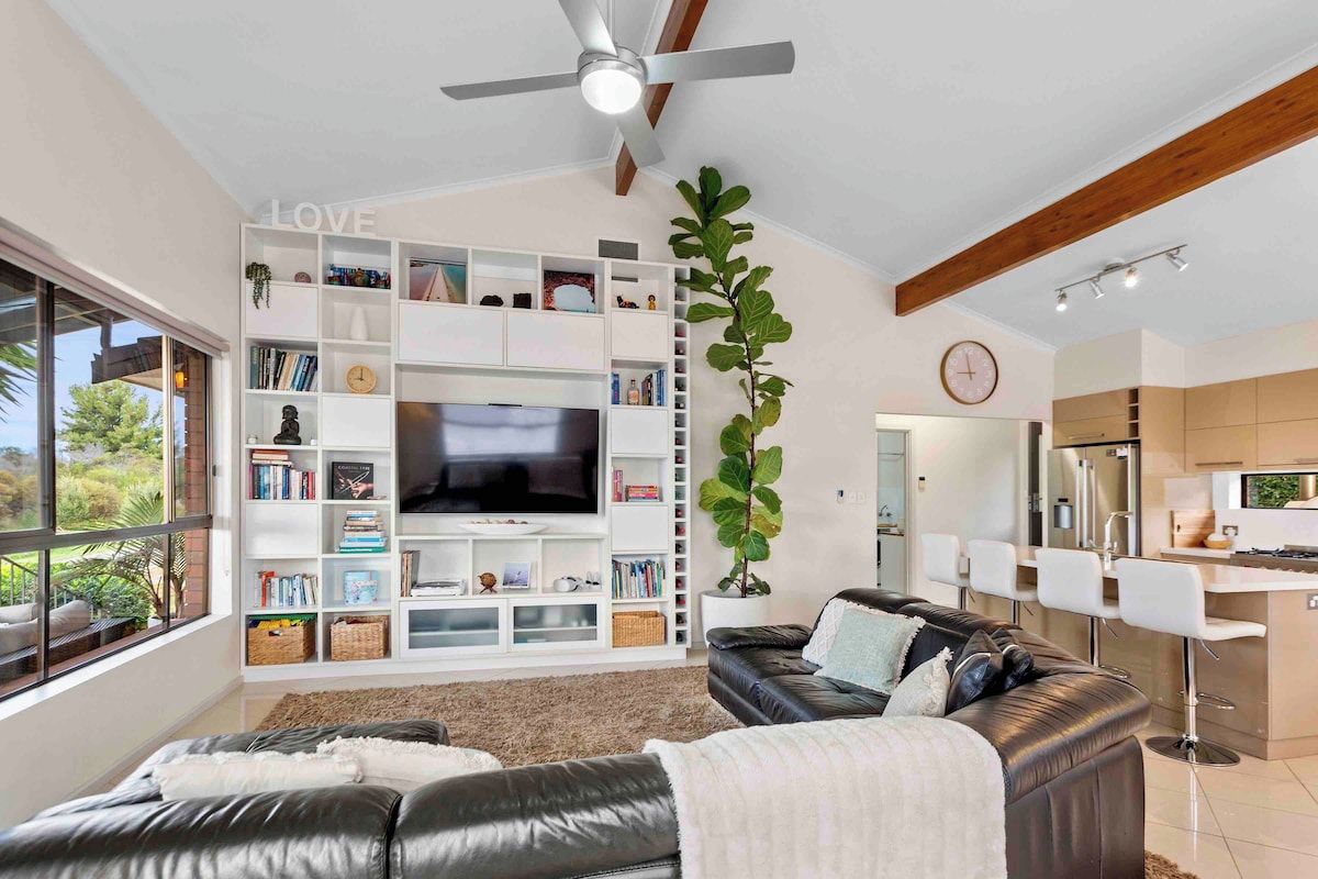 A modern living room with a flat-screen TV, a bookshelf, a large plant, a leather sofa, and a dining area with bar stools.