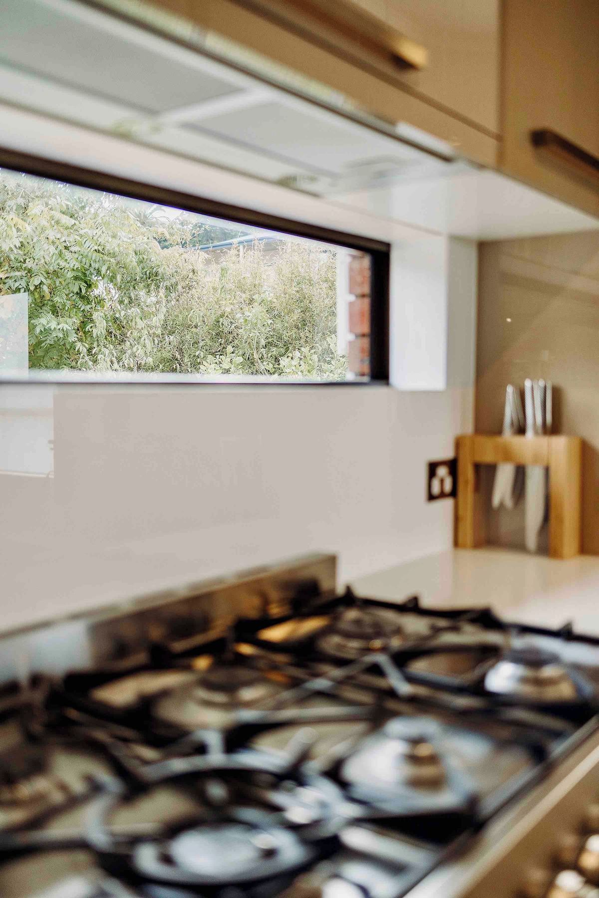 A kitchen with a gas stove, a window with a view of greenery, and a wooden cabinet with utensils.