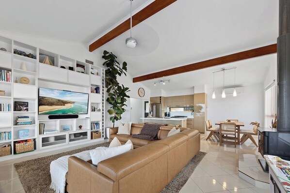 A modern living room with a large flat-screen TV, a sofa, a dining area with a table and chairs, and a kitchen in the background.