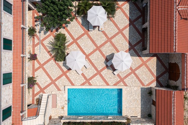 Vue aérienne d’un espace piscine sur le toit, avec une piscine à l’eau d’un bleu limpide, entourée d’un sol recouvert de carreaux rouges et de parasols blancs.