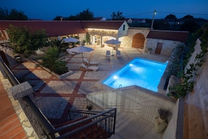 A swimming pool with lounge chairs and umbrellas, stone flooring, and a stone building with a red roof in the background.