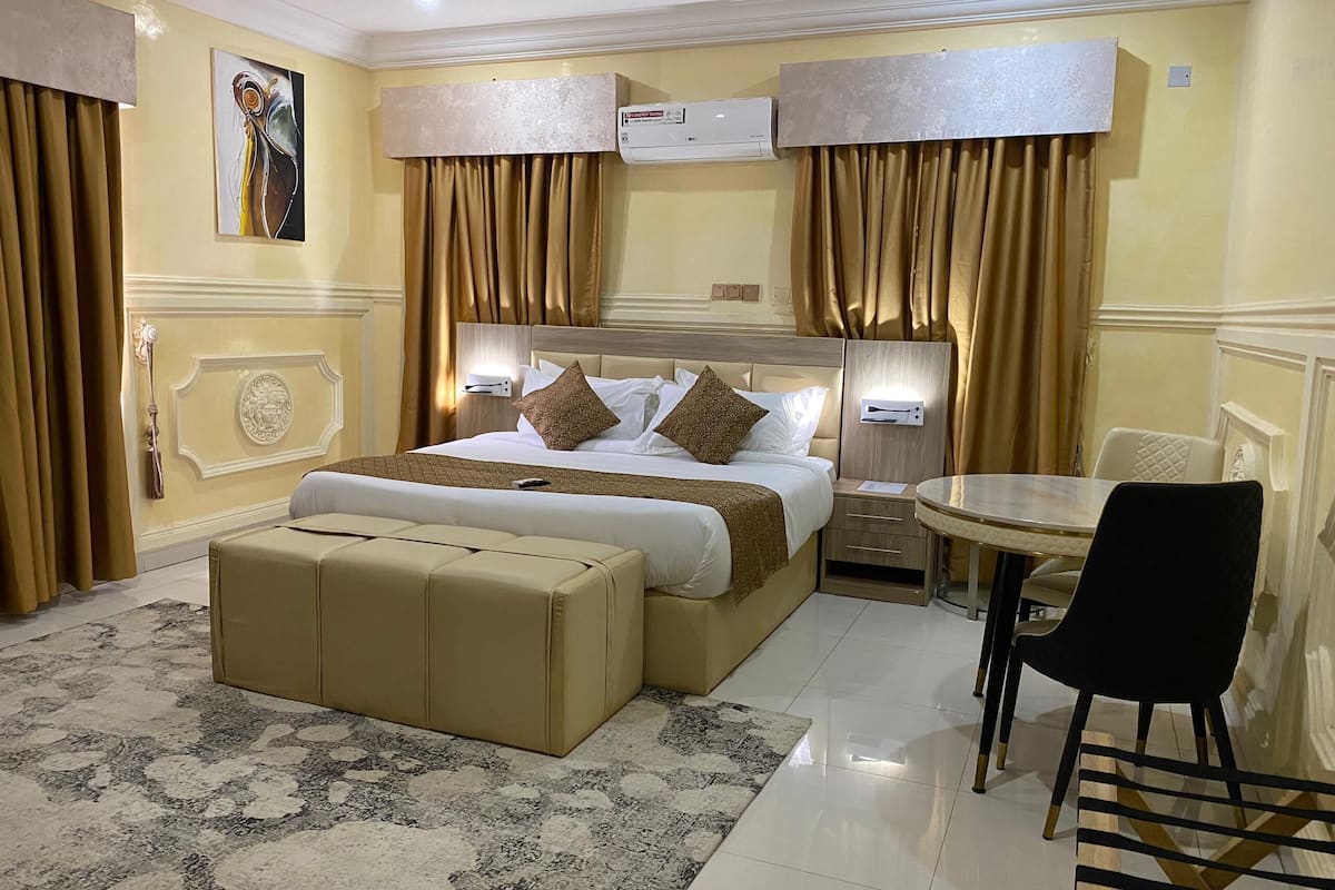 A hotel room with a large bed, a bedside table, a chair, a small round table, and a wall-mounted air conditioner.