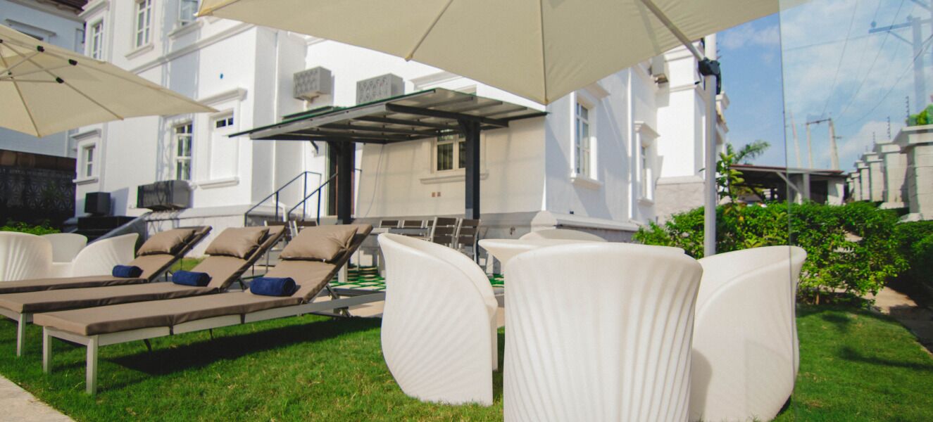 Zona de piscina con sillones de descanso y sombrillas, un área de estar techada y un edificio al fondo.