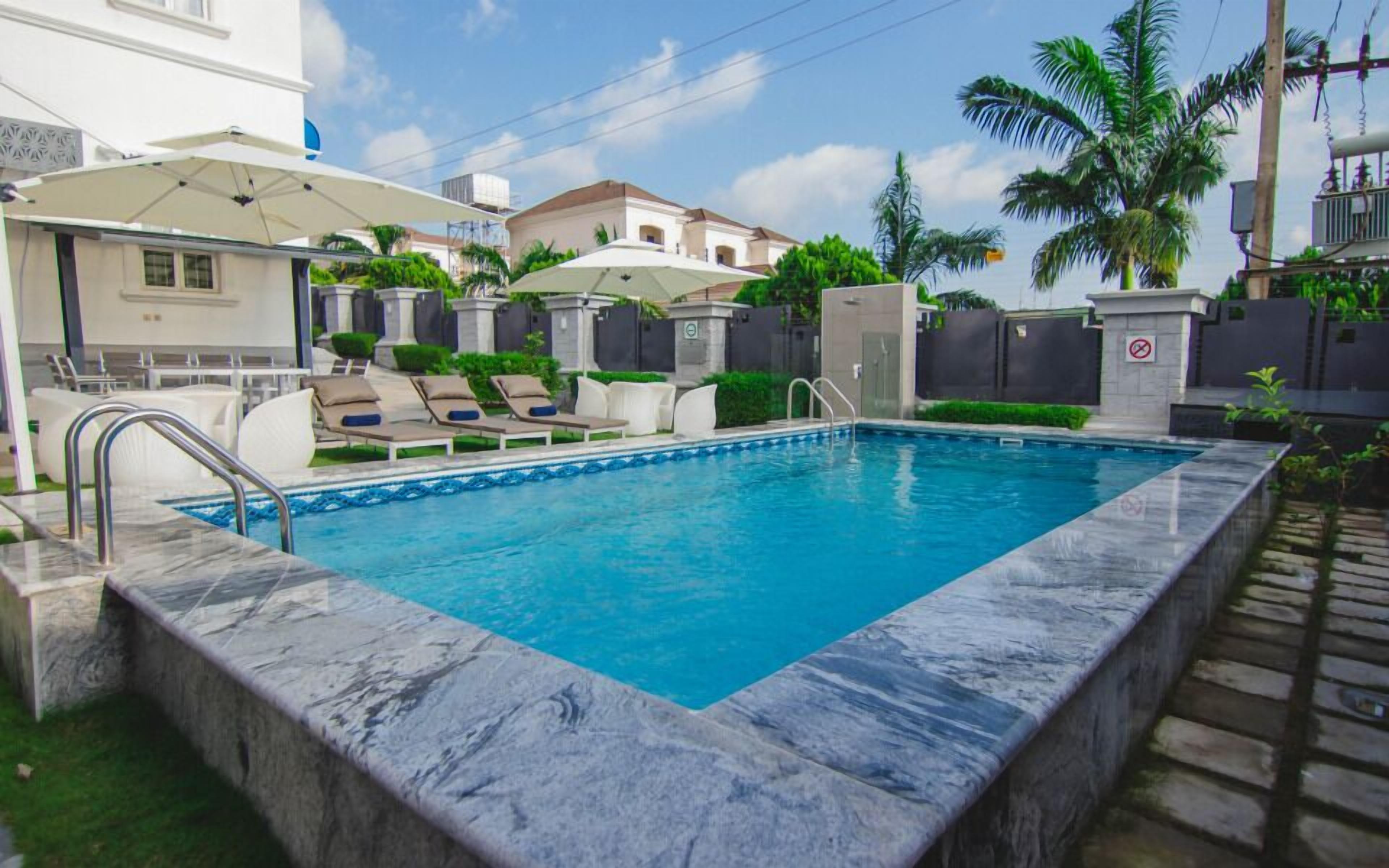 Una piscina rectangular con borde de mármol, rodeada de sillas de descanso y sombrillas, con vistas a edificios residenciales y palmeras al fondo.