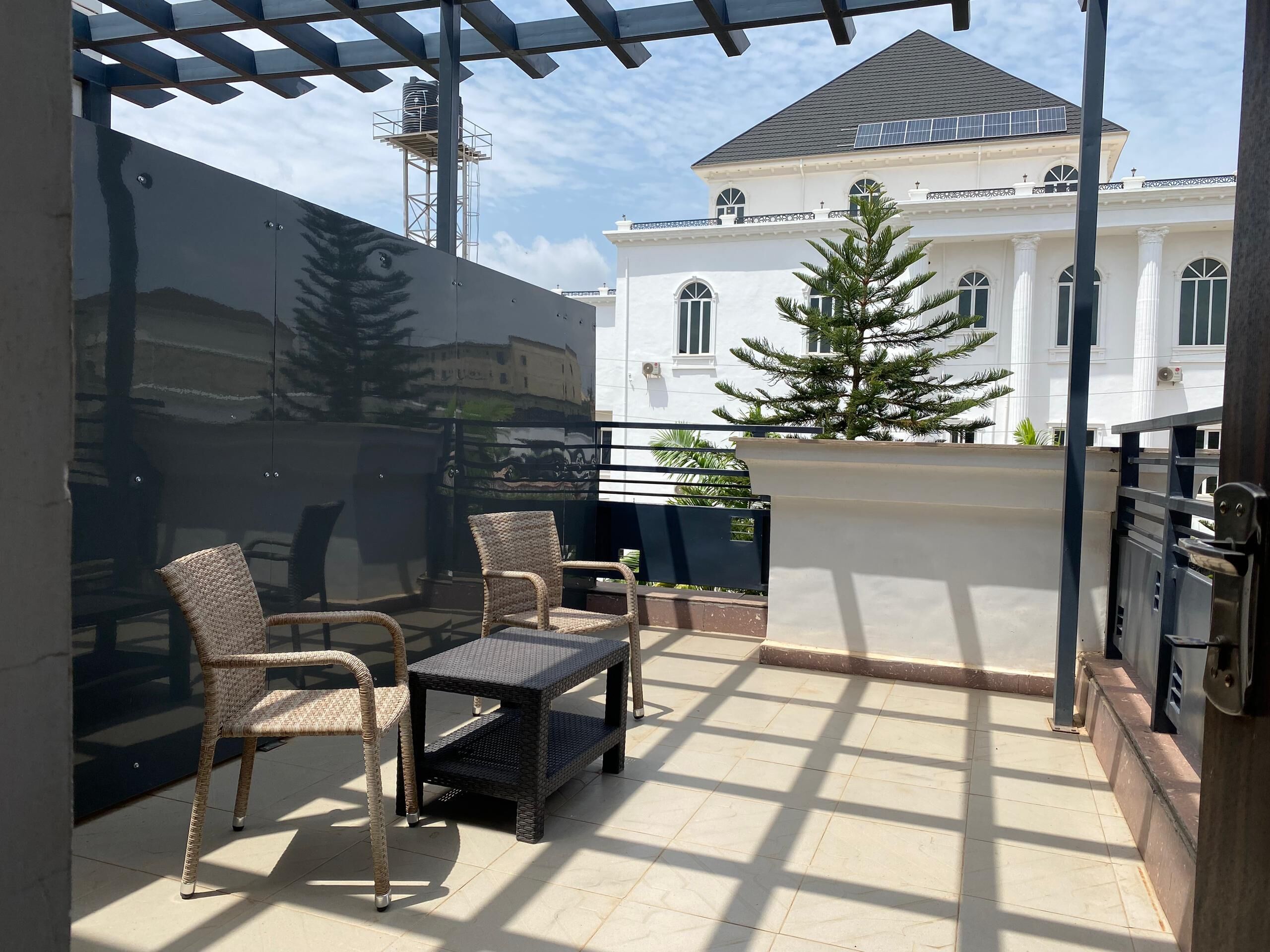 Une terrasse extérieure aménagée avec des meubles en osier, une petite table et une vue sur un bâtiment blanc doté de panneaux solaires.