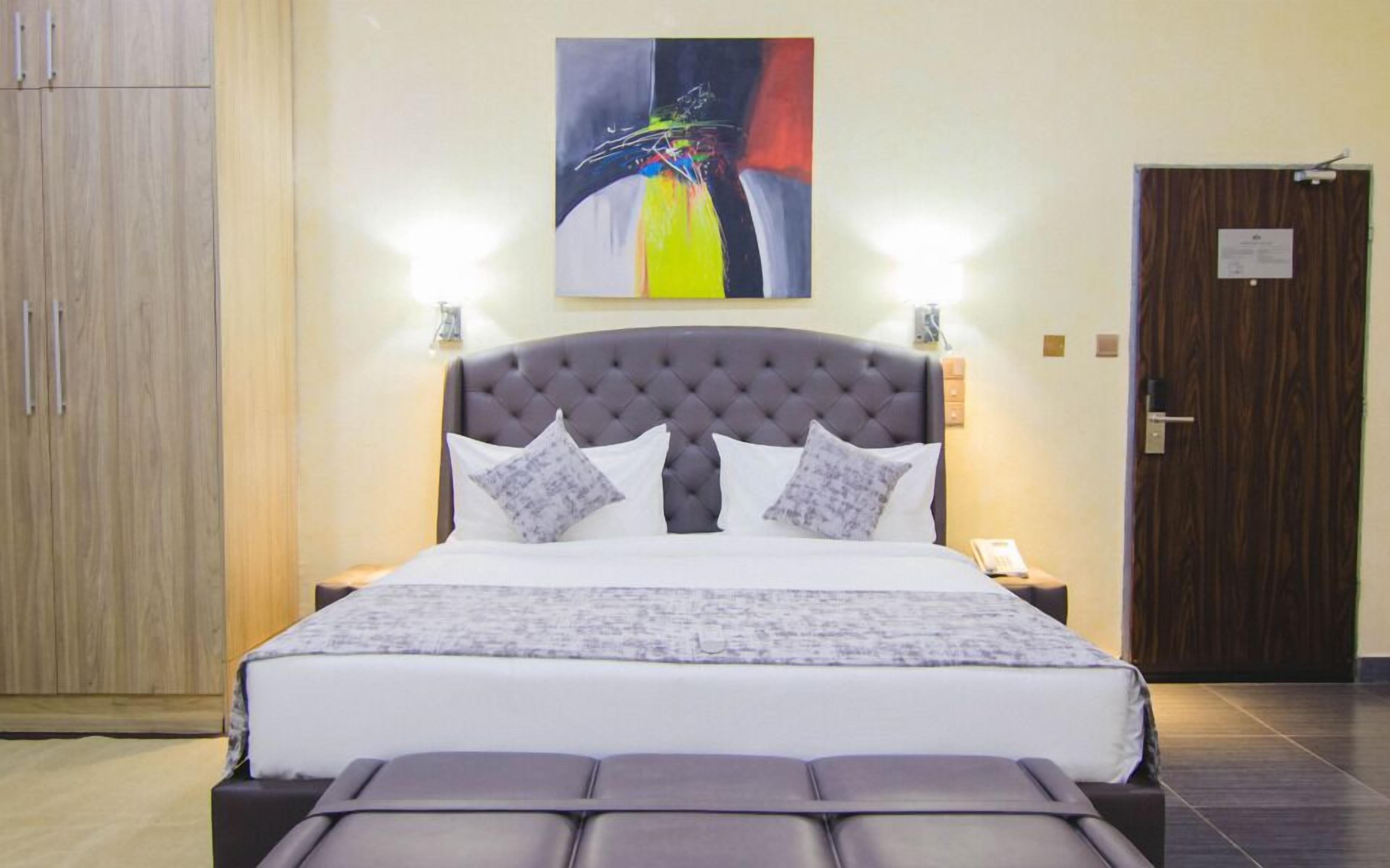 A hotel room with a large bed, a grey upholstered headboard, and a colorful abstract painting above the bed.