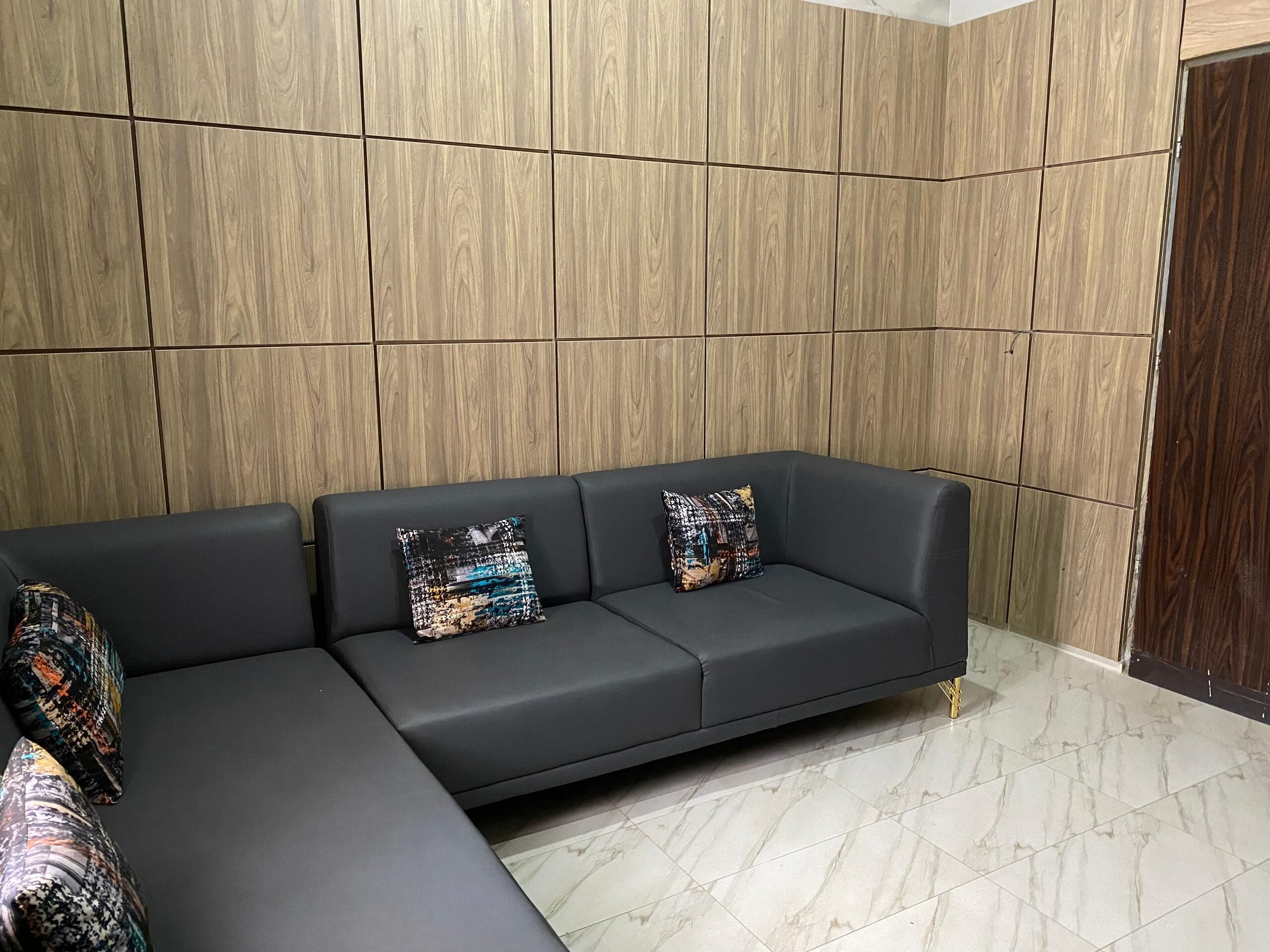 A modern living area with a dark-colored sofa and patterned cushions, set against a wooden panel wall and a tiled floor.