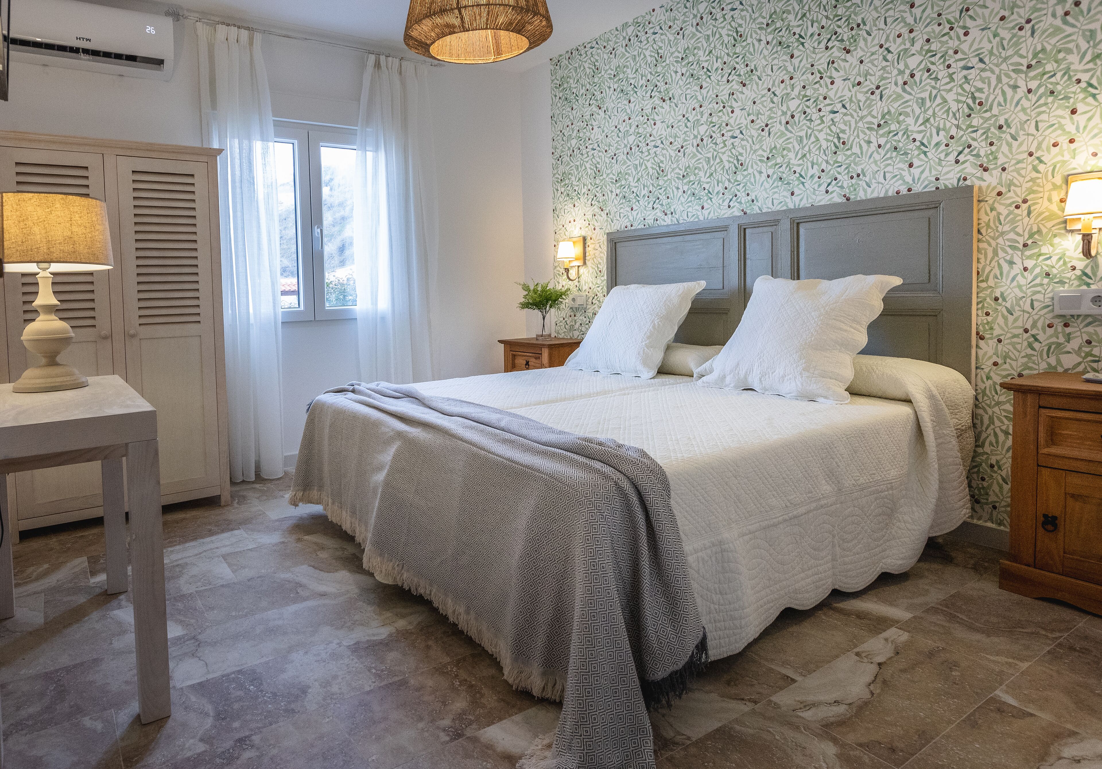 Un dormitorio ordenado con cama, mesitas de noche, lámpara y ventana con cortinas.