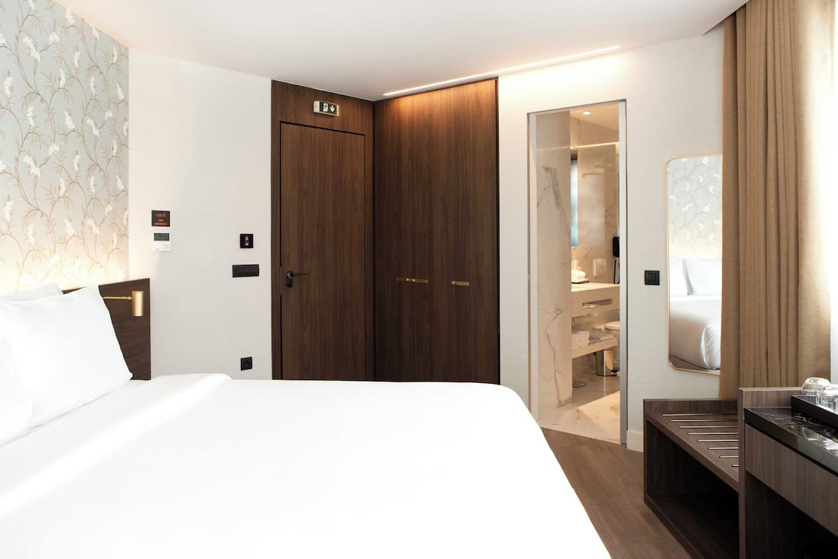 Una habitación de hotel moderna con una cama grande, un armario de madera, un vestidor y un baño con espejo.