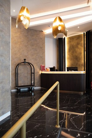 A modern reception area with a marble floor, a reception desk, a luggage cart, and two pendant lights. - Luwian Athens Boutique Hotel (Athens)