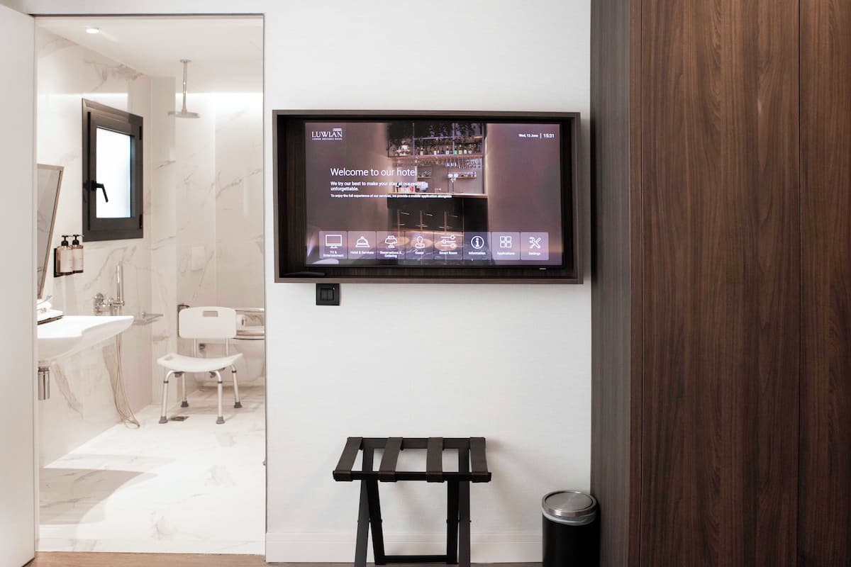 Habitación de hotel moderna con una pantalla grande mostrando un mensaje de bienvenida, un banco de madera y un cesto de basura.