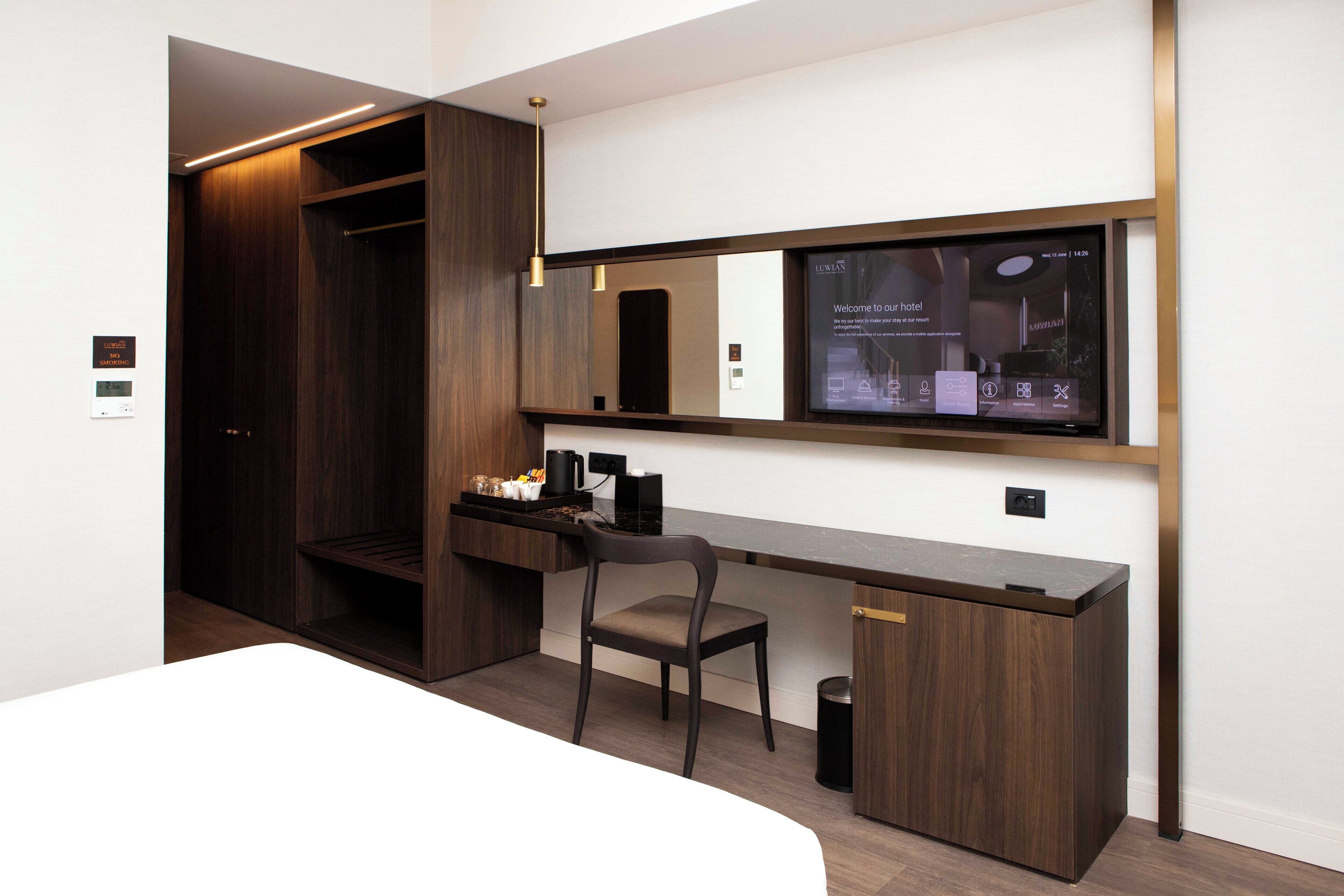 A modern hotel room with a wooden desk, a chair, a large mirror, and a digital display on the wall.