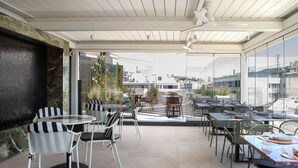 A rooftop terrace with tables and chairs, large windows, and a view of the cityscape.