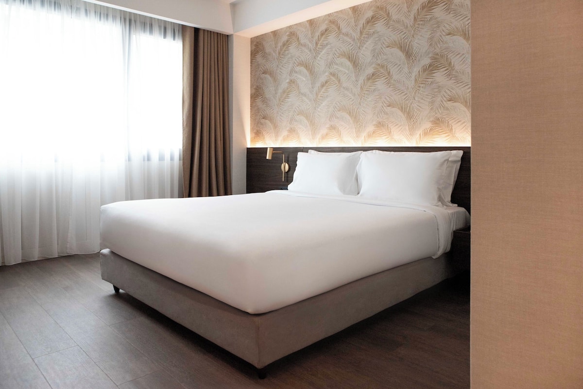 Una habitación de hotel moderna con una cama grande, un cabecero estampado y vistas al exterior a través de cortinas traslúcidas.