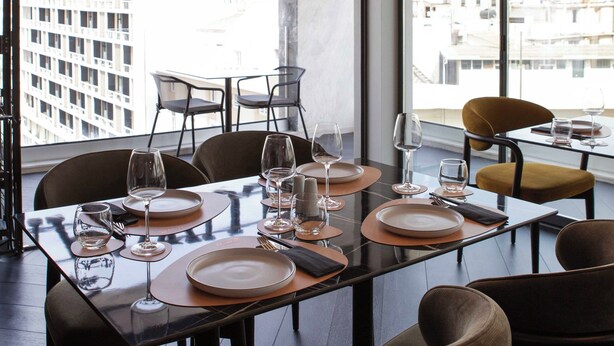 A dining area with a table set for six, large windows with a city view, and modern lighting fixtures.