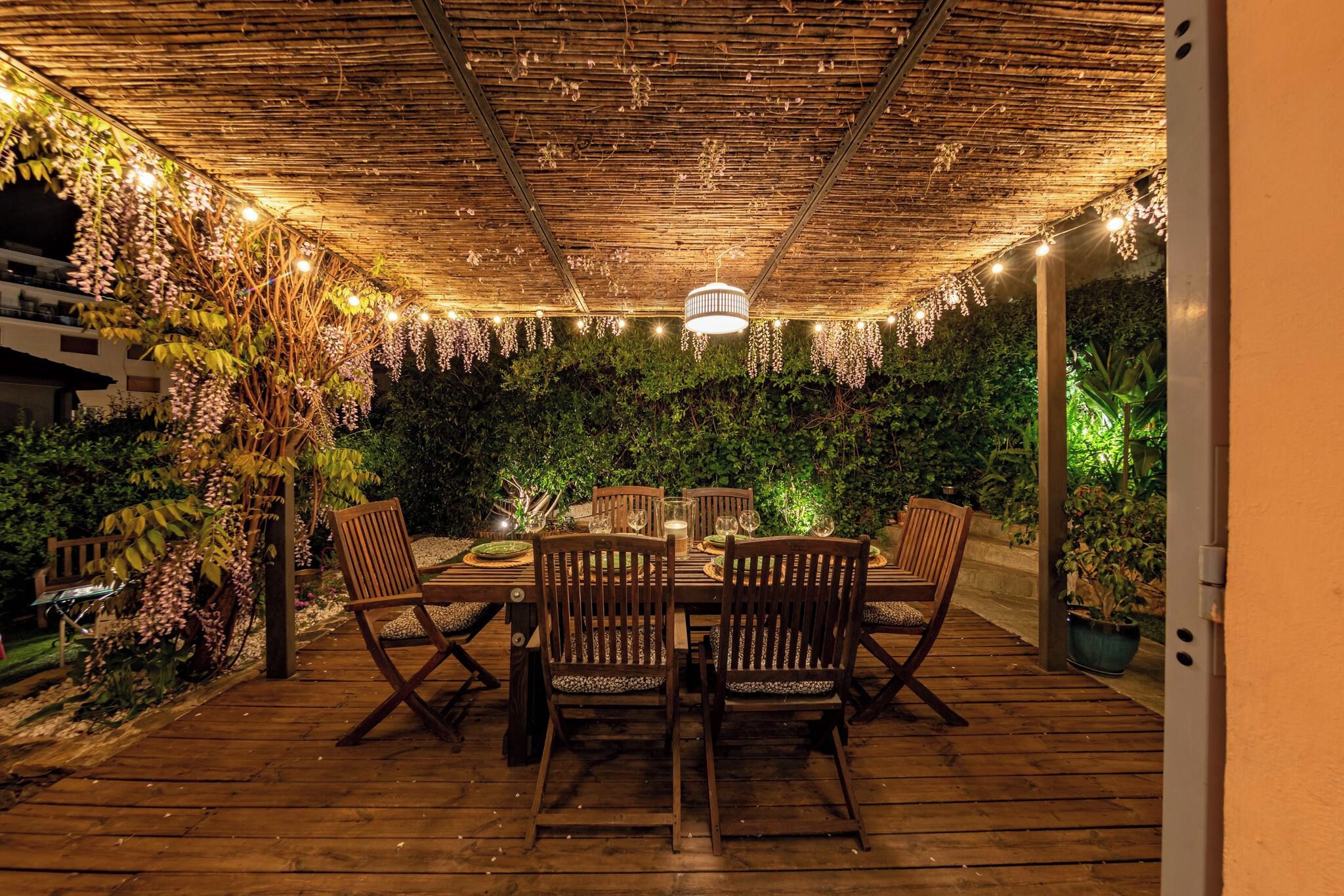 Un área de comedor al aire libre con mesa y sillas de madera, rodeada de abundante follaje e iluminación colgante.